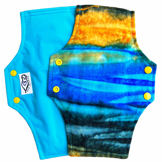 Two reusable menstrual pads with squared top and flared bottom shape against a white background. The pad on top is made from flannel fabric with batik striations of yellow, aqua and indigo. The pad on the bottom is made from aqua blue PUL fabric.