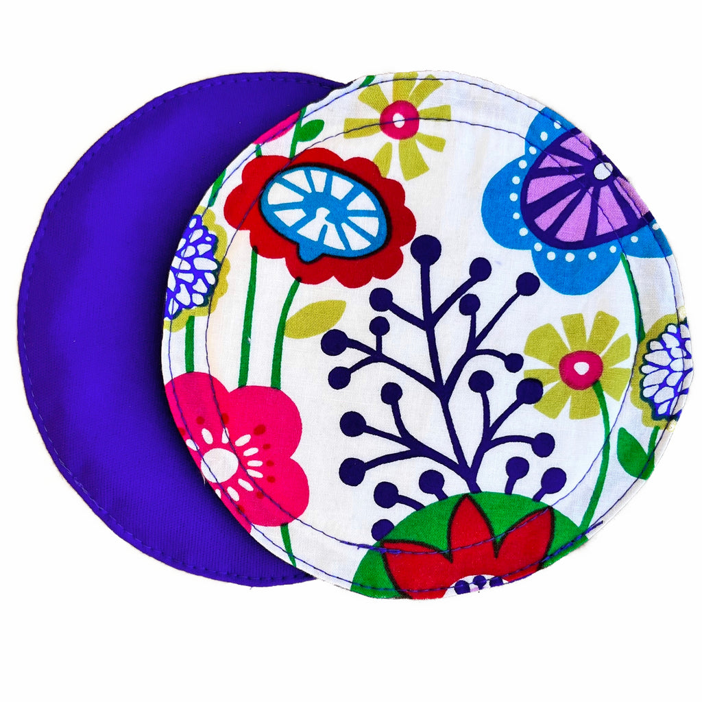 Two reusable nursing pads against a white background. The breast pad on top is made from white woven cotton fabrics with a bright floral pattern. The breast pad on the bottom is made from grape purple PUL fabric.