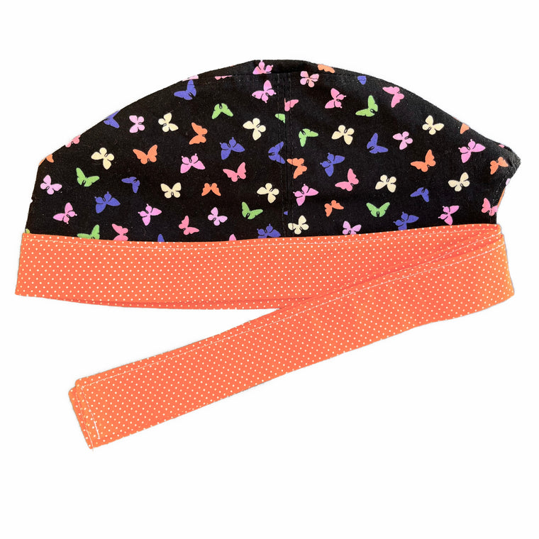A black and orange scrub cap against a white background. The main part of the cap is made from black woven cotton with mulitcoloured pastel butterflies. The bottom band and ties are made from orange woven cotton with very small white dots.