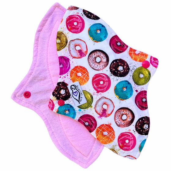 Two reusable menstrual pads with a contoured shape, against a white background. The pad on top is made from white cotton fabric with small multicoloured donuts print, and has a white "Scarlet Eve" label. The pad on the bottom is made from light pink flannel fabric.
