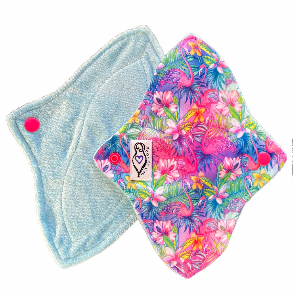 Two reusable menstrual pads with a contoured shape against a white background. The pad on top is made from  light blue fabric with a bright, tropical flamingos print and has a white "Scarlet Eve" label. The pad on the bottom is made from light blue bamboo velour fabric.