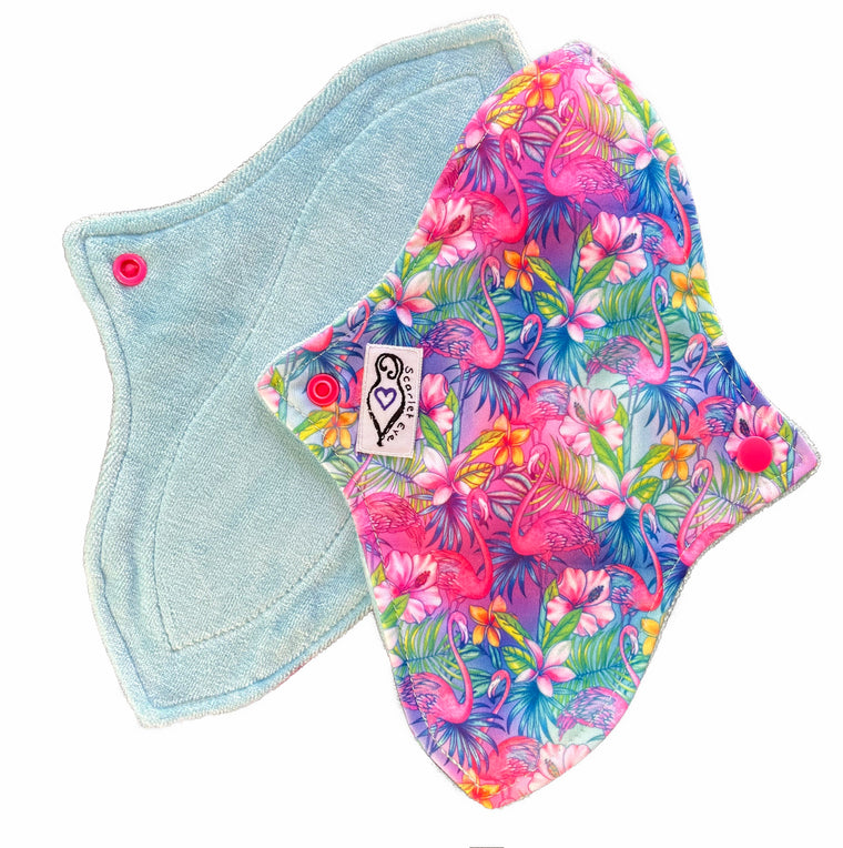 Two reusable menstrual pads with a contoured shape against a white background. The pad on top is made from  light blue fabric with a bright, tropical flamingos print and has a white "Scarlet Eve" label. The pad on the bottom is made from light blue bamboo velour fabric.