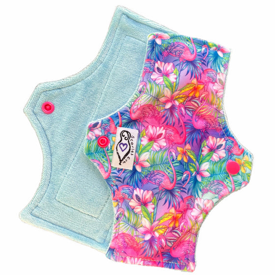 Two reusable menstrual pads with squared top and flared bottom shape against a white background. The pad on top is made from  light blue fabric with a bright, tropical flamingos print and has a white "Scarlet Eve" label. The pad on the bottom is made from light blue bamboo velour fabric.