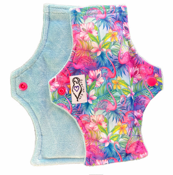 Two reusable menstrual pads with squared top and flared bottom shape against a white background. The pad on top is made from  light blue fabric with a bright, tropical flamingos print and has a white "Scarlet Eve" label. The pad on the bottom is made from light blue bamboo velour fabric.