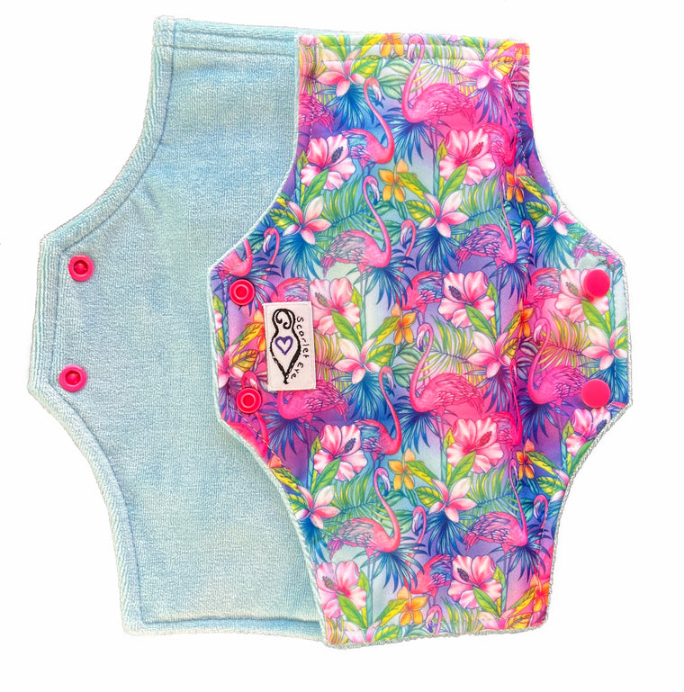 Two reusable menstrual pads with squared top and flared bottom shape against a white background. The pad on top is made from  light blue fabric with a bright, tropical flamingos print and has a white "Scarlet Eve" label. The pad on the bottom is made from light blue bamboo velour fabric.
