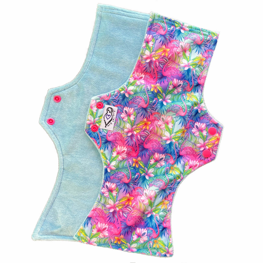 Two reusable menstrual pads with squared top and flared bottom shape against a white background. The pad on top is made from light blue PUL fabric with a bright, tropical flamingos print and has a white "Scarlet Eve" label. The pad on the bottom is made from light blue bamboo velour fabric.
