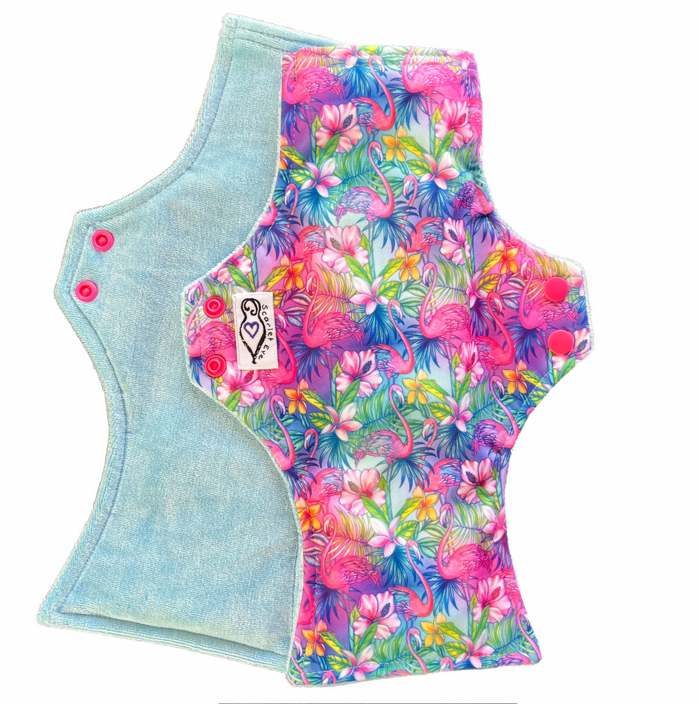 Two reusable menstrual pads with squared top and flared bottom shape against a white background. The pad on top is made from  light blue fabric with a bright, tropical flamingos print and has a white "Scarlet Eve" label. The pad on the bottom is made from light blue bamboo velour fabric.