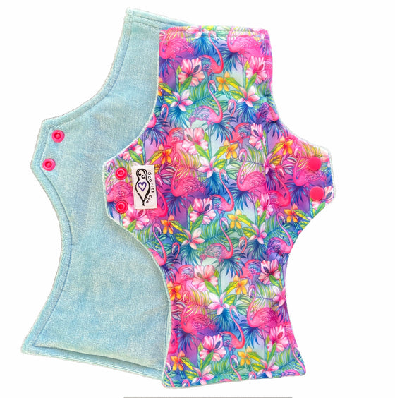 Two reusable menstrual pads with squared top and flared bottom shape against a white background. The pad on top is made from  light blue fabric with a bright, tropical flamingos print and has a white "Scarlet Eve" label. The pad on the bottom is made from light blue bamboo velour fabric.