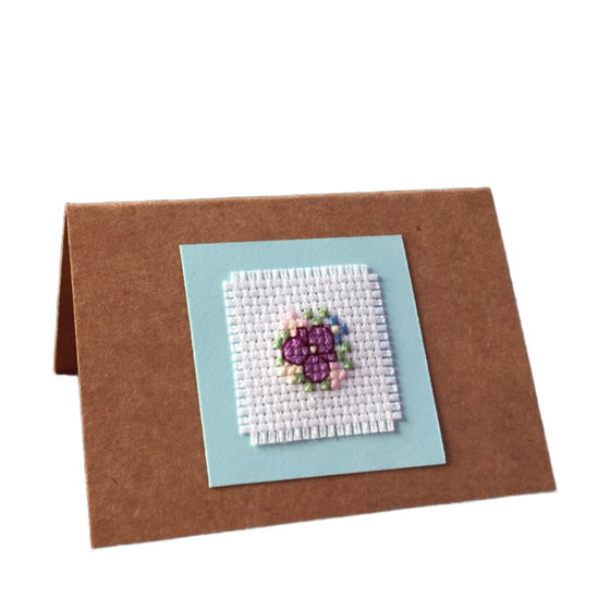 A handcrafted cardboard gift tag with a cross stitch design featuring a floral pattern in colors purple, green, and blue, mounted on a brown card.
