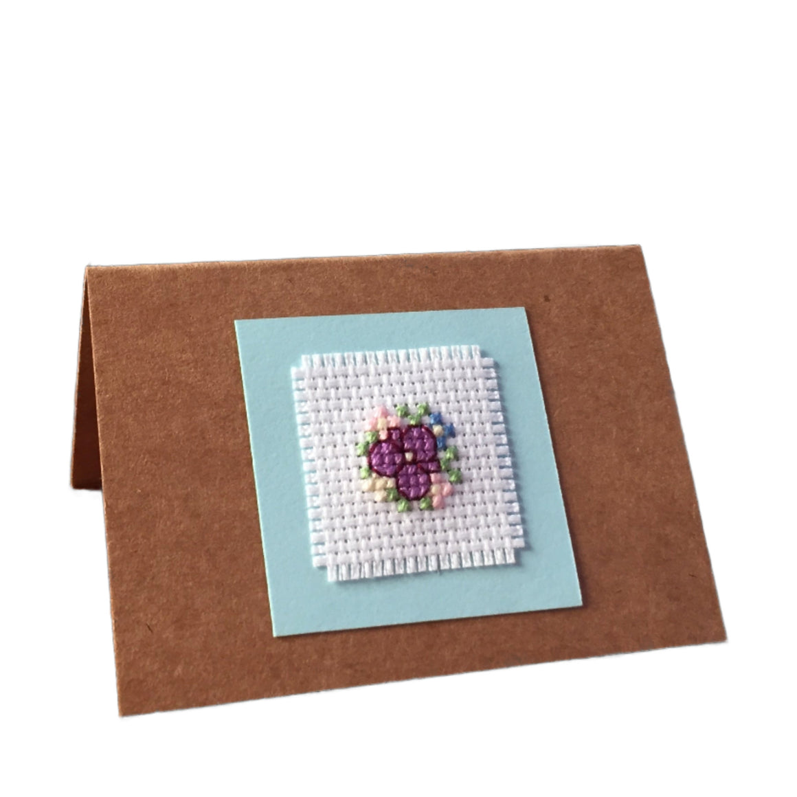 A handcrafted cardboard gift tag with a cross stitch design featuring a floral pattern in colors purple, green, and blue, mounted on a brown card.