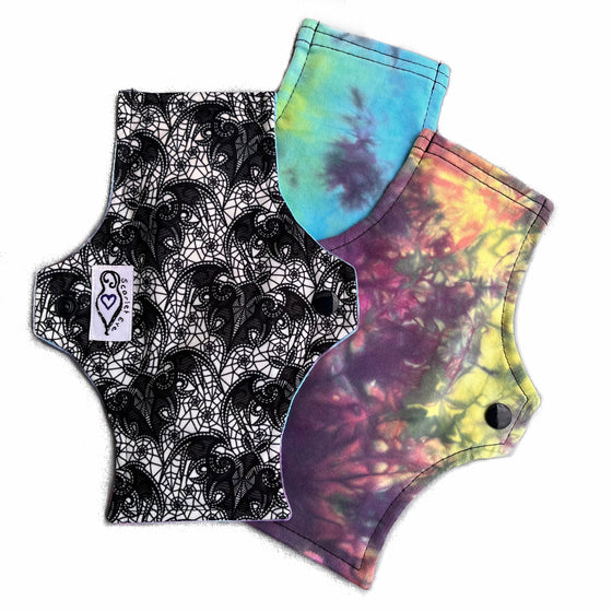 Three reusable cloth menstrual pads with a squared top and flared bottom shape, against a white background. The pad on the top is made from white PUL fabric with a black lace bats print, and has a white "Scarlet Eve" label. The pads on the bottom are made from tie dye cotton lycra fabric in pastel and black shades.