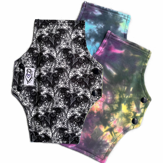 Three reusable cloth menstrual pads with a squared top and flared bottom shape, against a white background. The pad on the top is made from white PUL fabric with a black lace bats print, and has a white "Scarlet Eve" label. The pads on the bottom are made from tie dye cotton lycra fabric in pastel and black shades.