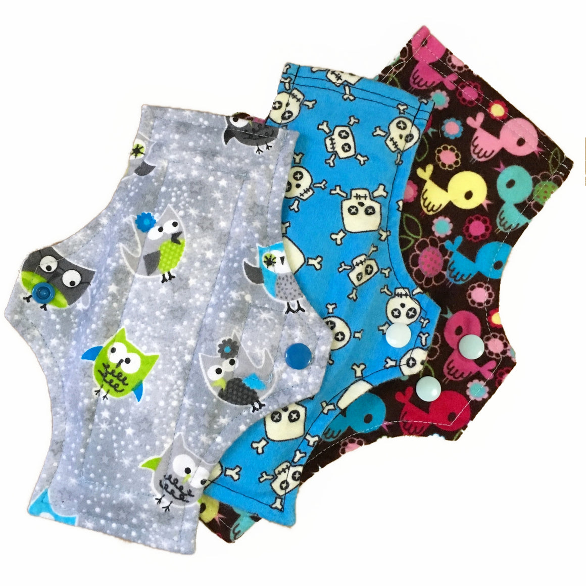 Three reusable menstrual pads with squared top and flared bottom shape against a white background. The Pads are arranged from largest to smallest. The pad on top is made from grey cotton flannel with grey, green and blue owls. The pad in the middle is made from aqua blue cotton flannel with black and white skulls. The pad on the bottom is made from brown cotton flannel with muliticoloured birds.