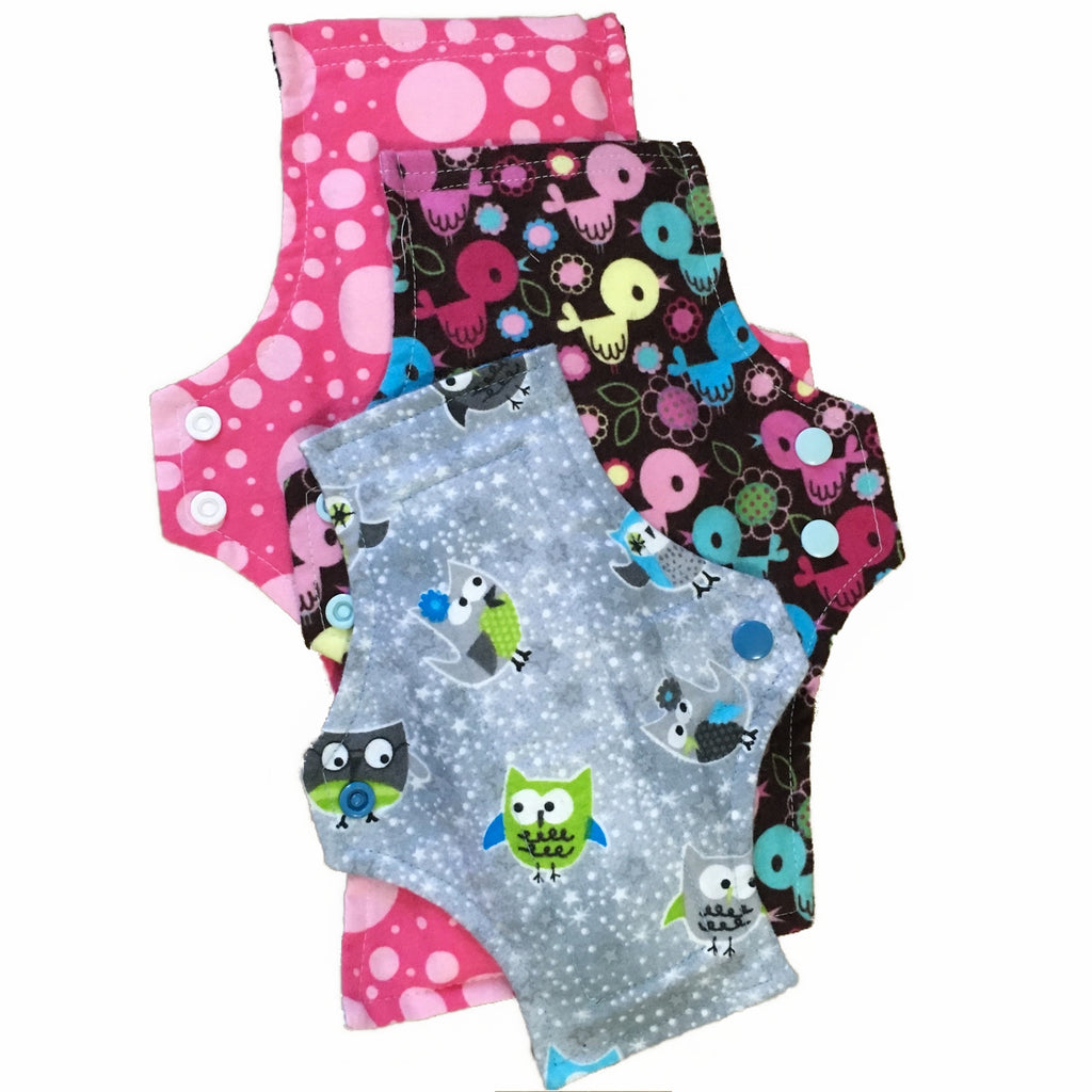 Three reusable menstrual pads with squared top and flared bottom shape against a white background. The Pads are arranged from largest to smallest. The pad on top is made from grey cotton flannel with grey, green and blue owls. The pad in the middle is made from brown cotton flannel with muliticoloured birds. The pad on the bottom is made from pink cotton flannel with light pink circles.