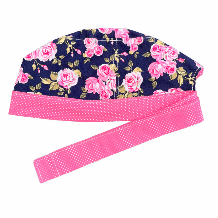 A navy and pink scrub cap against a white background. The main part of the cap is made from navy woven cotton with pink roses. The bottom band and ties are made from pink woven cotton with very small white dots.