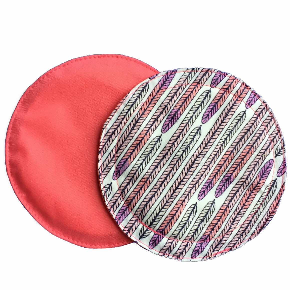 Two reusable nursing pads against a white background. The breast pad on top is made from cream woven cotton fabric with pink and violet feathers. The breast pad on the bottom is made from coral pink PUL fabric.