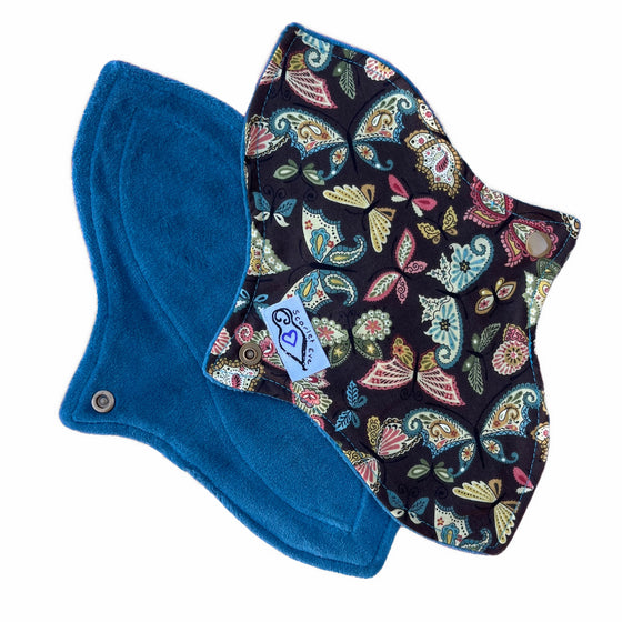 Two reusable menstrual pads with a contoured shape, against a white background. The pad on top is made from dark brown cotton fabric with pink and blue paisley butterflies print, and has a white "Scarlet Eve" label. The pad on the bottom is made from jade green velour fabric.