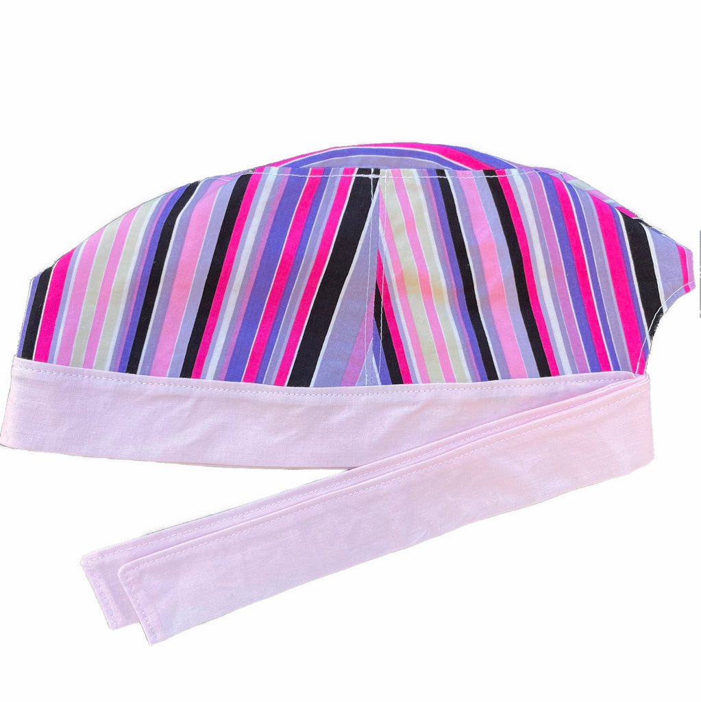 A pink and purple scrub cap against a white background. The main part of the cap is made from woven cotton with purple, pink, black and light yellow stripes. The bottom band and ties are made from light pink woven cotton.