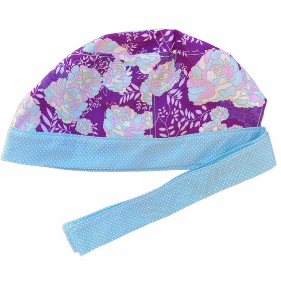 A purple and blue scrub cap against a white background. The main part of the cap is made from plum purple woven cotton with pastel blue, pink, and yellow floral print. The bottom band and ties are made from light blue woven cotton with very small white dots.