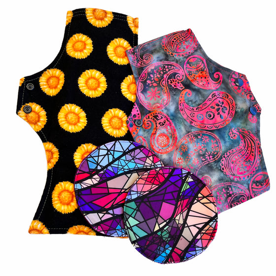 Two reusable menstrual pads with squared top and flared bottom shape and two reusable breast pads, against a white background. The larger pad is made from black flannel with bright yellow sunflowers and the smaller pad is made from denim blue woven cotton with a candy pink paisley pattern. The breast pads are made from a multicoloured stained glass print.