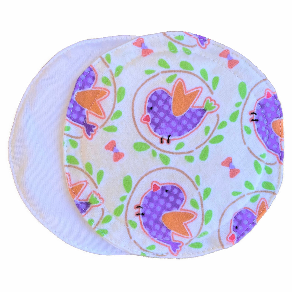 Two reusable nursing pads against a white background. The breast pad on top is made from cream flannel with purple and orange birds and green leaves. The breast pad on the bottom is made from white PUL fabric.