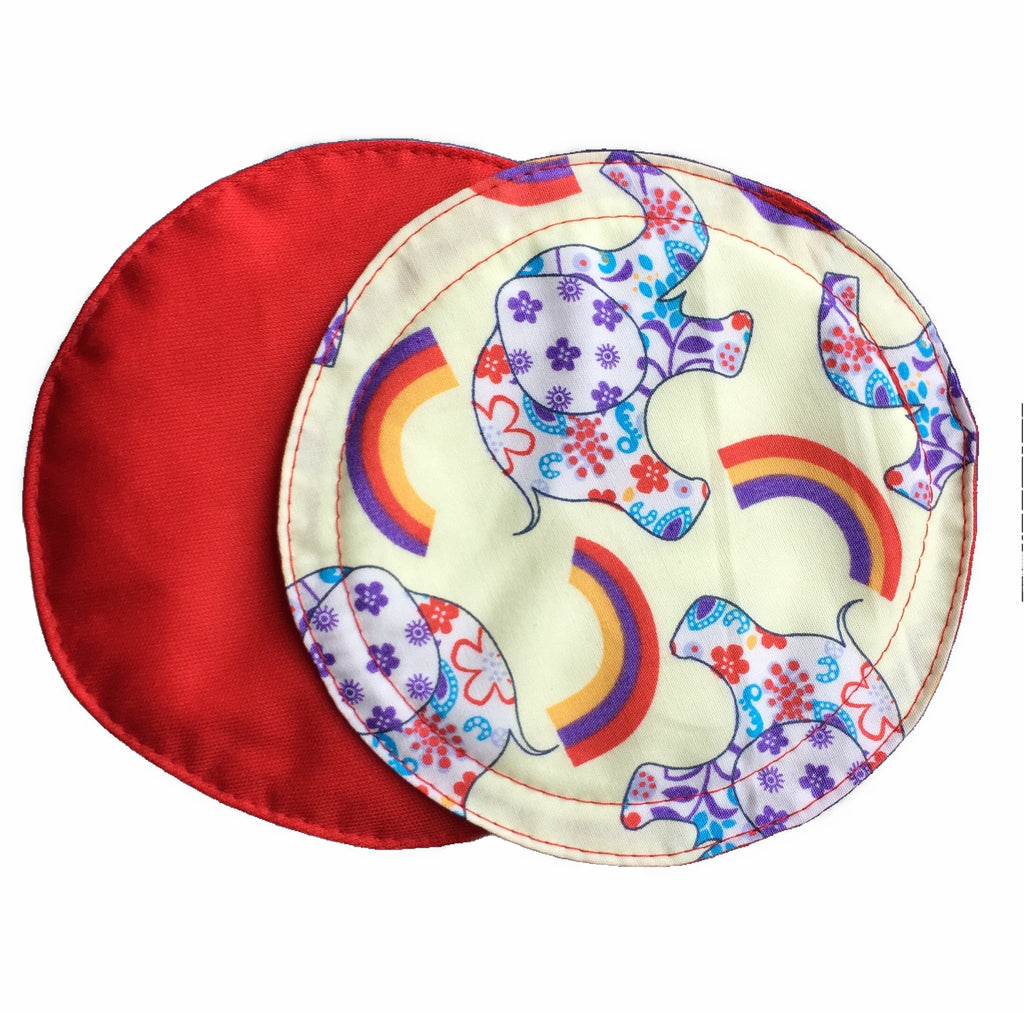 Two reusable nursing pads against a white background. The breast pad on top is made from light yellow woven cotton fabric with elephants and rainbows. The breast pad on the bottom is made from red PUL fabric.