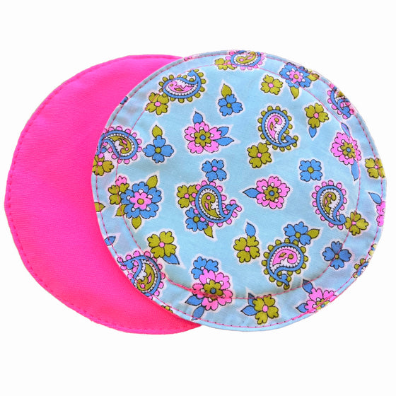 Two reusable nursing pads against a white background. The breast pad on top is made from light blue woven cotton with blue, pink and green floral paisley print. The breast pad on the bottom is made from neon pink PUL fabric.