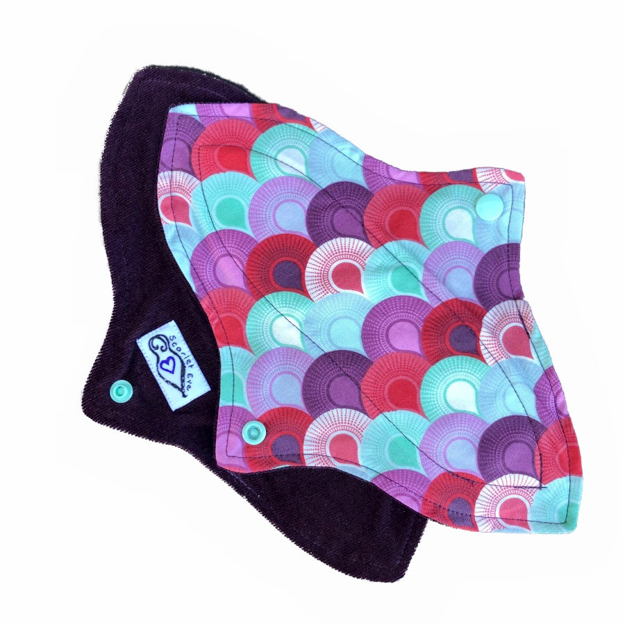 Two reusable menstrual pads with a contoured shape against a white background. The pad on top is made from woven cotton with purple, aqua and red rising sun shapes. The pad on the bottom is made from purple corduroy fabric and has a white "Scarlet Eve" label.
