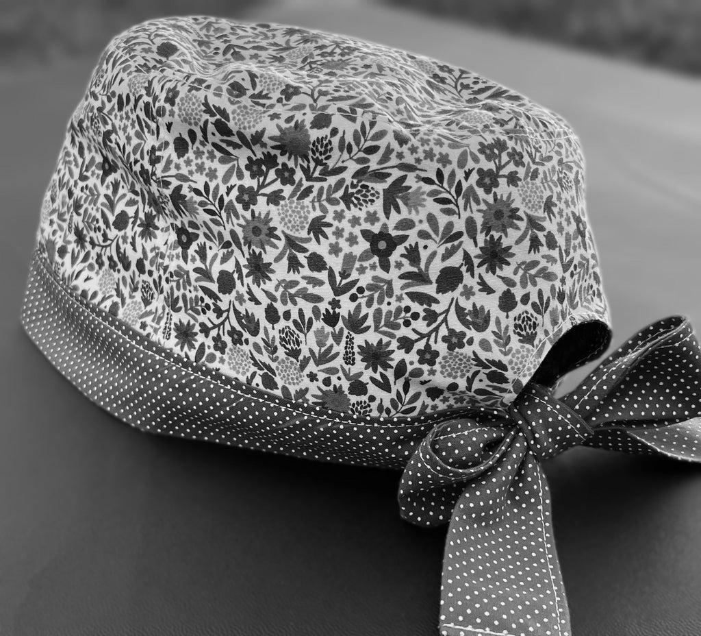 A black and white photo of a scrub cap with ties in a bow, against a solid grey background. The main part of the cap is floral, and the bottom band and ties are small white dots on a darker base.