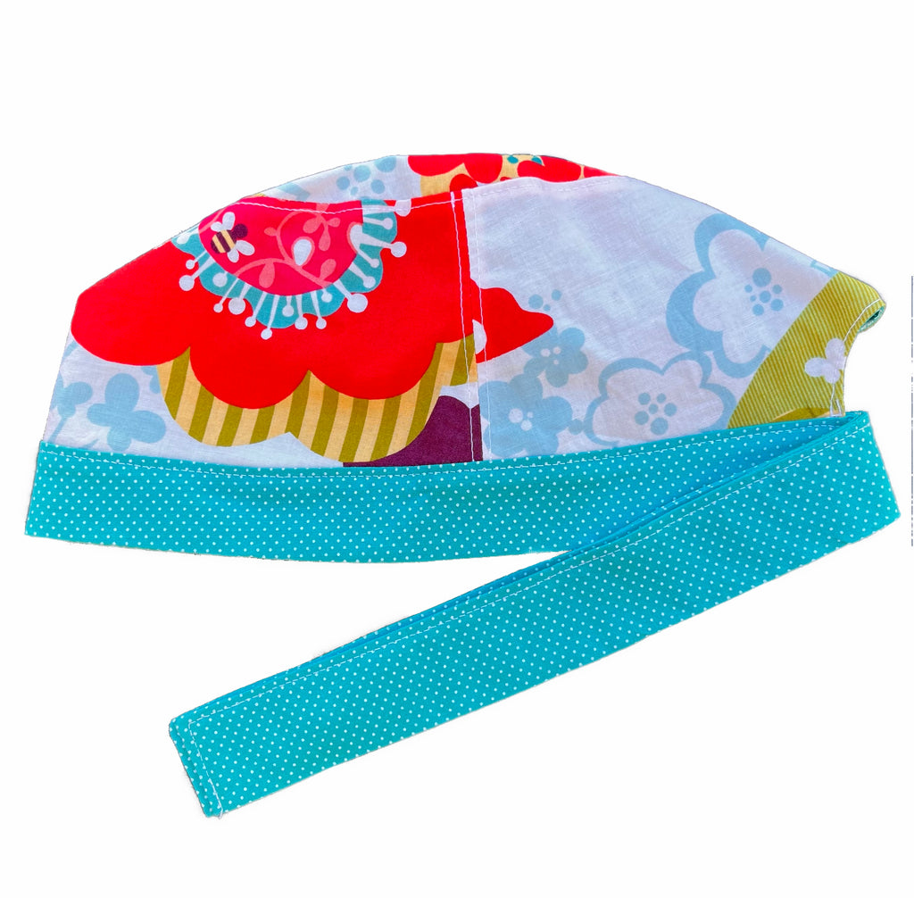 A bright print and turquoise scrub cap against a white background. The main part of the cap is made from white woven cotton with a large, bright floral print. The bottom band and ties are made from turquoise woven cotton with very small white dots.