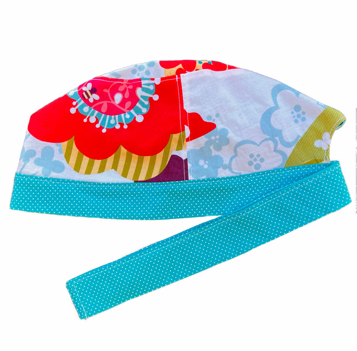 A bright print and turquoise scrub cap against a white background. The main part of the cap is made from white woven cotton with a large, bright floral print. The bottom band and ties are made from turquoise woven cotton with very small white dots.