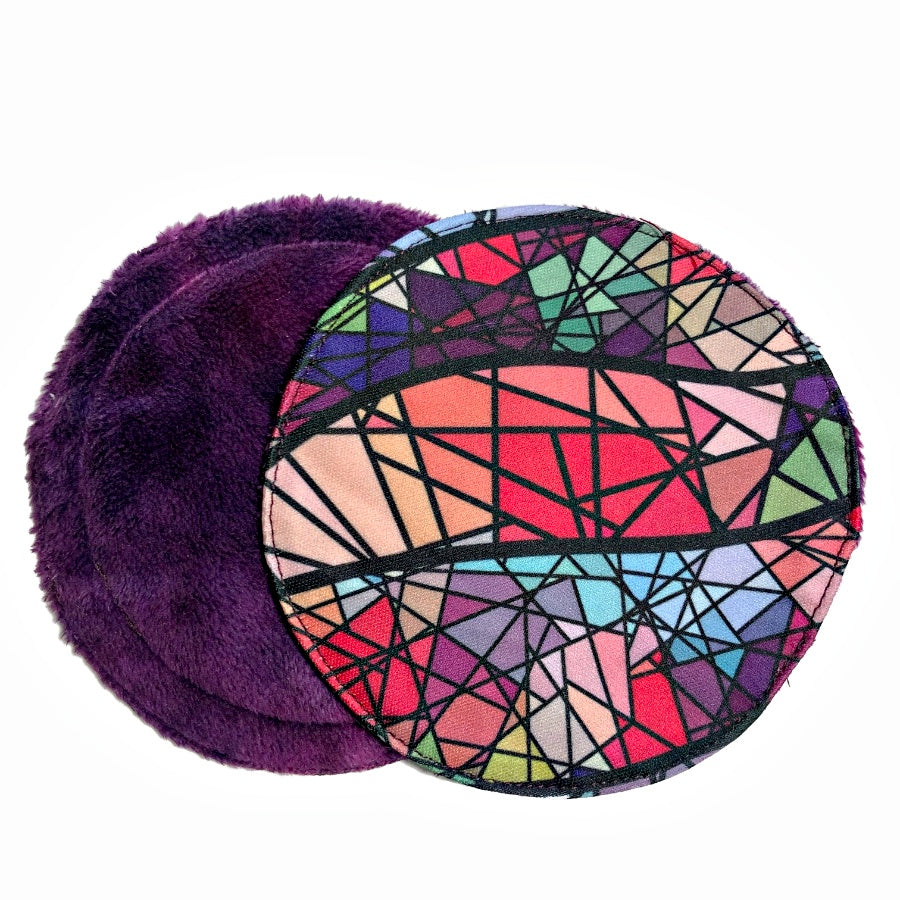 Two reusable nursing pads against a white background. The breast pad on top is made from PUL fabric with multicoloured triangles inside thick black lines. The breast pad on the bottom is made from purple minky fabric.