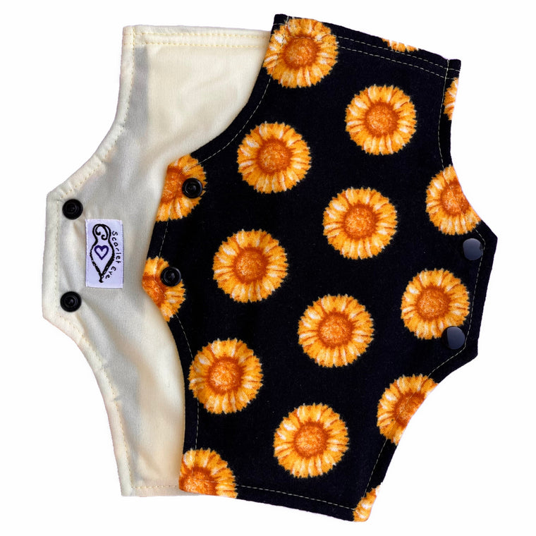 Two resusable menstrual pads with a squared top and flared bottom shape, against a white background. The pad on top is made from black flannel fabric with yellow sunflowers print. The pad underneath is made from pale yellow PUL fabric, and has a white "Scarlet Eve" label.