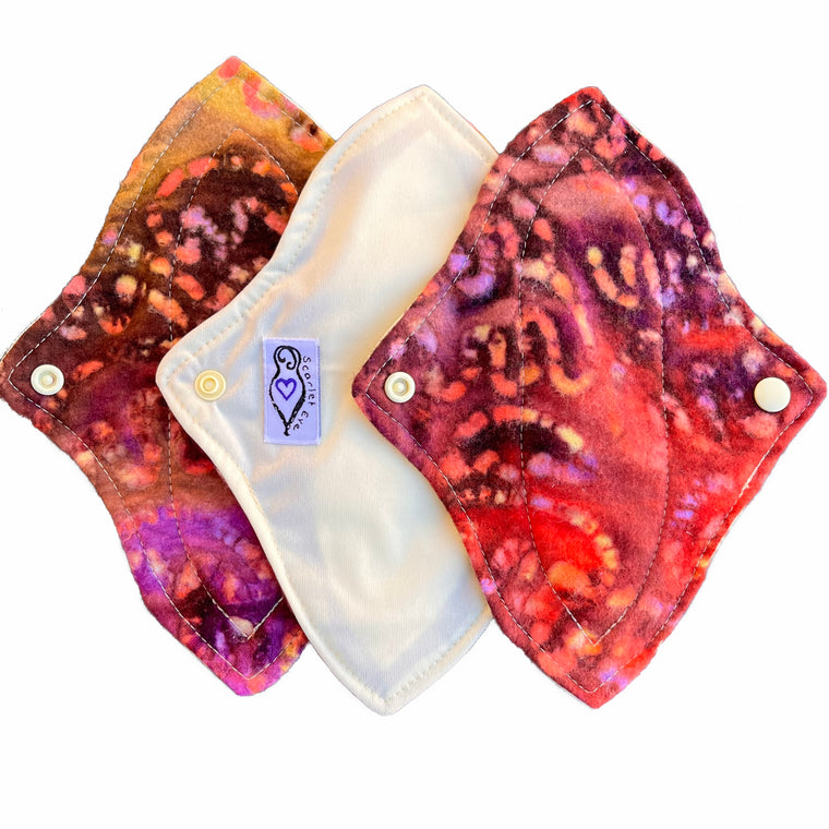 Three reusable cloth menstrual pads with a contoured shape, against a white background. The pad on the left is made from brown and purple batik flannel fabric. The pad in the middle is made from pale yellow PUL fabric, and has a white "Scarlet Eve" label. The pad on the left is made from red and purple batik flannel fabric.