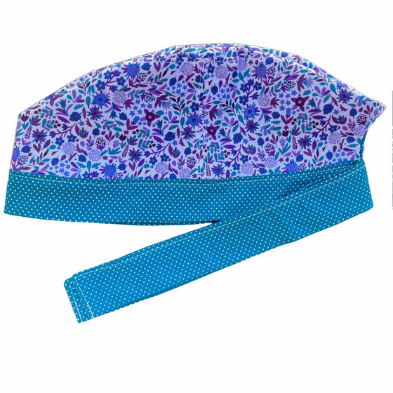 A teal and white scrub cap against a white background. The main part of the cap is made from white woven cotton with blue, purple and teal floral print. The bottom band and ties are made from teal woven cotton with very small white dots.
