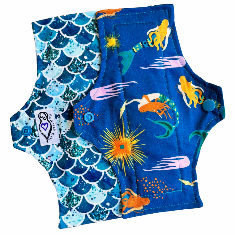 Two reusable menstrual pads with a squared top and flared bottom shape, against a white background. The pad on top is made from teal woven cotton with swimming mermaids. The pad on the bottom is made from PUL fabric with mermaid scales in various shades of green, and has a white "Scarlet Eve" label.