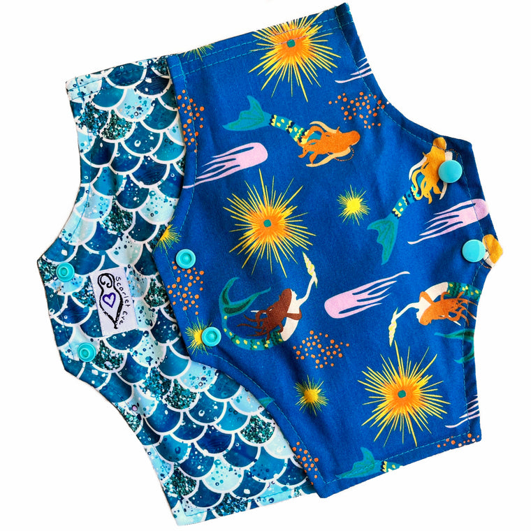 Two reusable menstrual pads with a squared top and flared bottom shape, against a white background. The pad on top is made from teal woven cotton with swimming mermaids. The pad on the bottom is made from PUL fabric with mermaid scales in various shades of green, and has a white "Scarlet Eve" label.