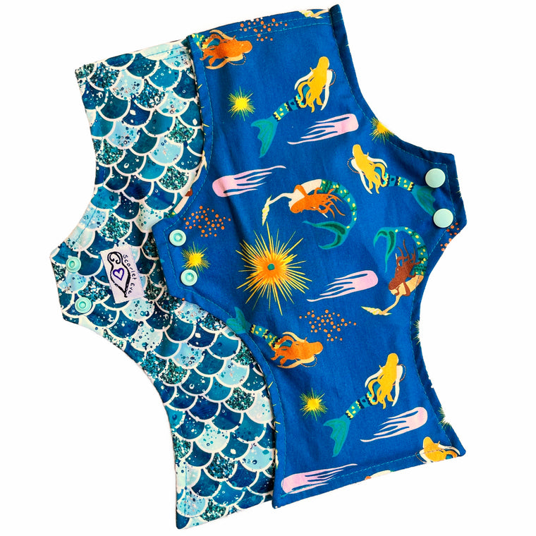 Two reusable menstrual pads with a squared top and flared bottom shape, against a white background. The pad on top is made from teal woven cotton with swimming mermaids. The pad on the bottom is made from PUL fabric with mermaid scales in various shades of green, and has a white "Scarlet Eve" label.