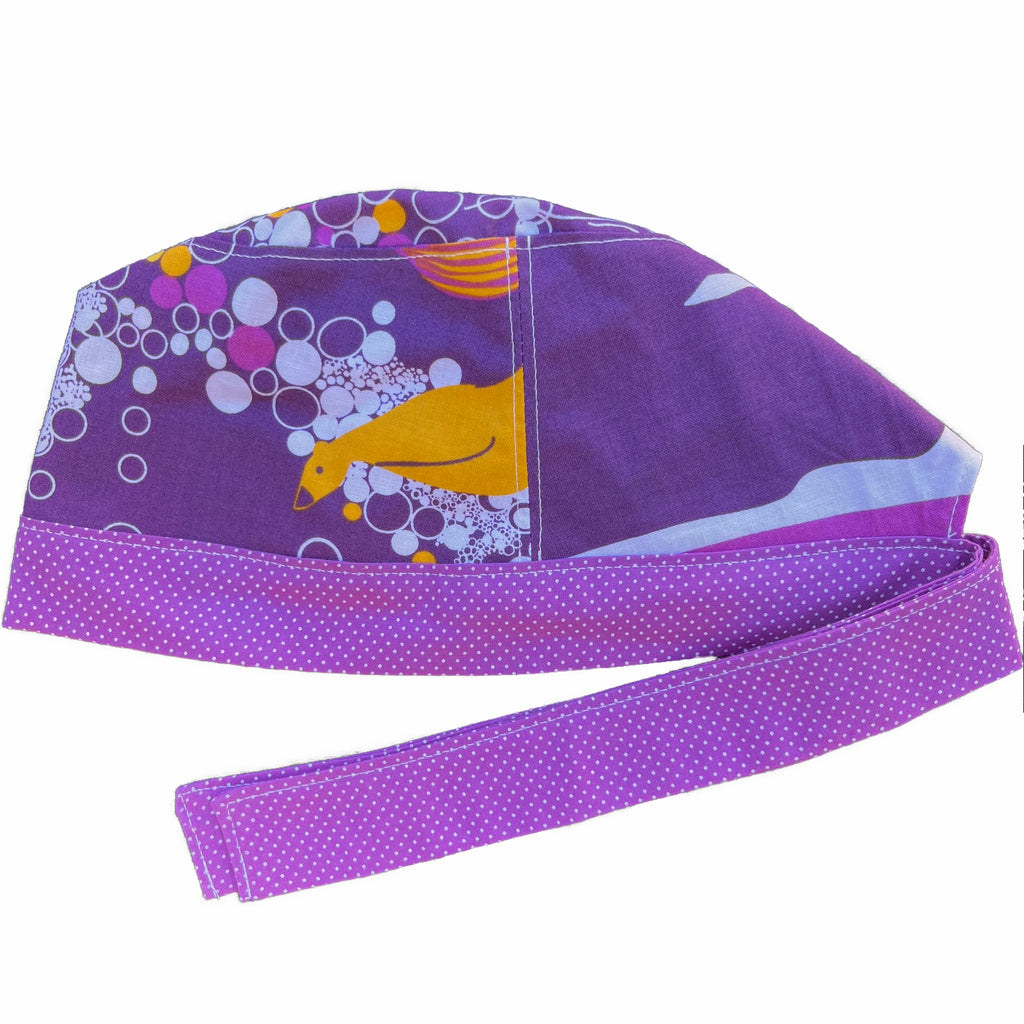 A violet and white scrub cap against a white background. The main part of the cap is made from violet woven cotton with yellow ducks and white circles. The bottom band and ties are made from violet woven cotton with very small white dots.