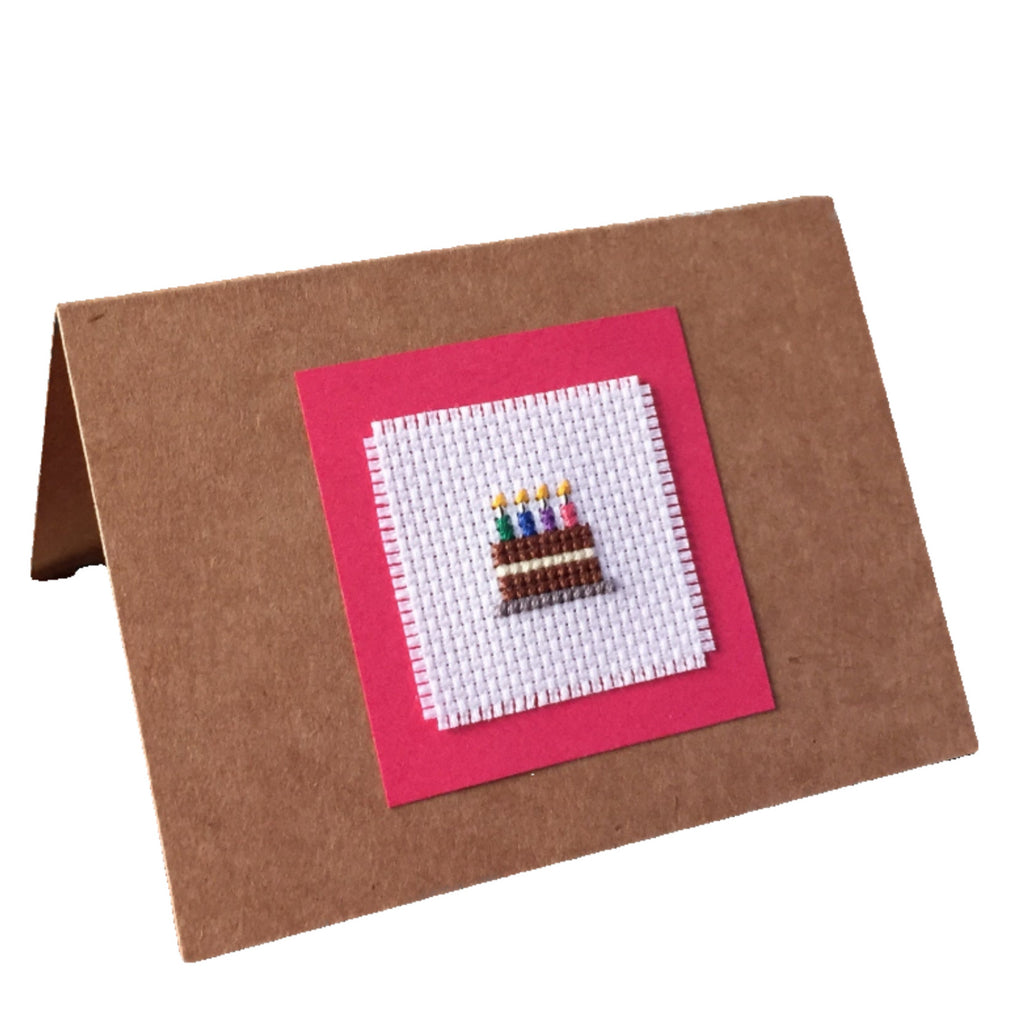 A handcrafted cardboard gift card with a cross stitch design featuring a birthday cake with candles, mounted on a brown card.