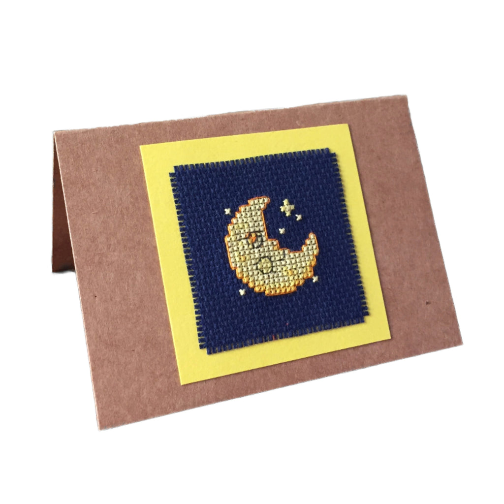 A handcrafted cardboard gift card with a cross stitch design featuring a yellow moon on a navy background, mounted on a brown card.